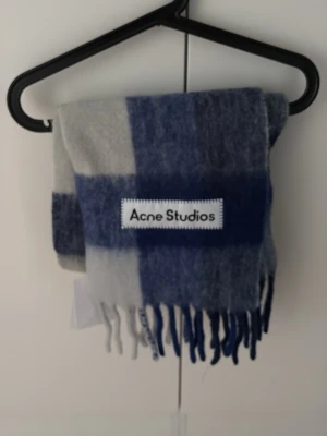 Blårutig halsduk från Acne Studios - Säljer en snygg halsduk från Acne Studios i blått och grått rutmönster. Halsduken har fransar i ändarna och är tillverkad i mjukt material, perfekt för att hålla värmen. Acne Studios-loggan är tydligt synlig på ena sidan.