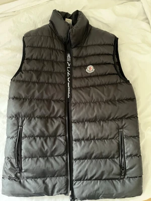Svart dunväst från Moncler - Snygg svart dunväst från Moncler med quiltad design och hög krage. Västen har två dragkedjefickor framtill och Moncler-logga på bröstet. Perfekt för lager-på-lager och ger en stilren look. Materialet är glansigt och fyllningen är dun.