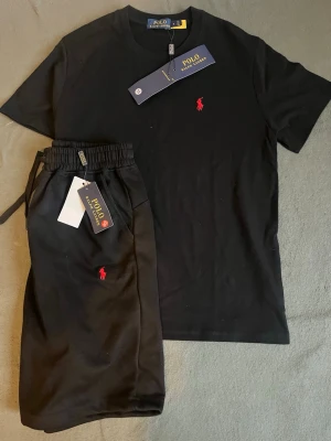 Svart set från Polo Ralph Lauren - Snyggt svart set från Polo Ralph Lauren med t-shirt och shorts. Båda plaggen har det klassiska röda Polo-logotrycket på bröstet och benet. T-shirten har rund hals och shortsen har dragsko i midjan. Materialet känns mjukt och bekvämt, perfekt för en avslappnad stil.