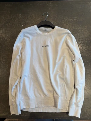 Vit sweatshirt från C.P. Company - Säljer en stilren vit sweatshirt från C.P. Company med rund hals och diskret logga broderad på bröstet. Tröjan har långa ärmar och är gjord i mjuk bomull för maximal komfort. Perfekt för en clean och avslappnad look.