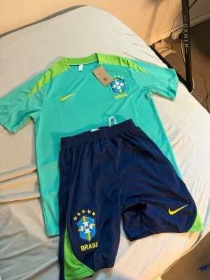 Brasilien fotbollsträningsset Nike - Säljer ett Brasilien fotbollsträningsset från Nike med turkos t-shirt och mörkblå shorts. Tröjan har limegröna detaljer och Brasilien-logga på bröstet. Shortsen har elastisk midja, Brasilien-logga och Nike swoosh. Perfekt för fotbollsträning eller match.