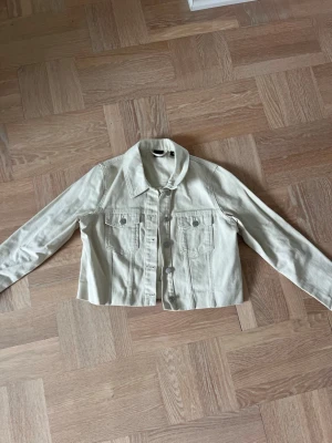 Ljusbeige jeansjacka från Vero Moda - Trendig ljusbeige jeansjacka från Vero Moda med klassisk krage, två bröstfickor med knappar och raka ärmar. Jackan har en croppad passform och stängs med knappar framtill. Perfekt att slänga över en t-shirt för en cool vardagslook.