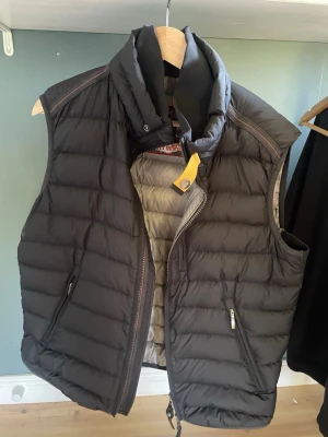 Svart dunväst från Parajumpers - Snygg svart dunväst från Parajumpers med hög krage och dragkedja framtill. Västen har två sidofickor med dragkedja och en cool gul detalj vid halsen. Insidan är ljusgrå och materialet är superlätt dun, perfekt för lager på lager. Skriv ifall ni har frågor. Några små fläckar, går säkert bort i tvätten. Kan skicka bild ifall de önskas.
