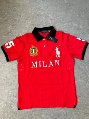 Polo Ralph Lauren Milan piké - Röd t-shirt från Polo Ralph Lauren med svart krage, vita broderade detaljer och MILAN-tryck på bröstet. Kortärmad modell i bomull med klubbemblem och stora siffror på ärmarna. Klassisk och sportig stil.