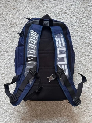 Mörkblå Nike ryggsäck med vita detaljer - Snygg mörkblå ryggsäck från Nike med vita detaljer och logga. Ryggsäcken har vadderade axelband med reflekterande mönster, flera fack och robust botten i svart material. Perfekt för skola eller träning och rymmer mycket.