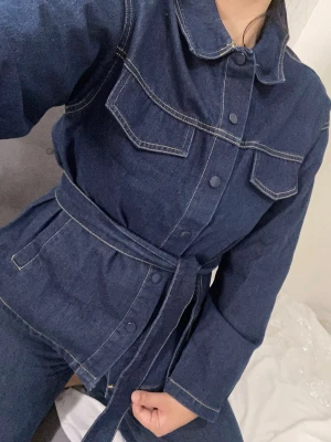 Mörkblå denim jeans set med bälte 50% bomull - Snygg mörkblå denim jeans set med sömmar, knappar framtill och två stora bröstfickor. Jumpsuiten har ett matchande tygbälte i midjan och långa ärmar för en cool och avslappnad look. Perfekt för dig som gillar jeansmaterial och vill ha en trendig helhet.