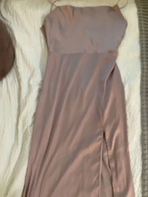 Lång beige/rosa klänning med slits - Säljer en elegant långklänning i beige/rosa med tunna axelband och en snygg slits framtill. Klänningen har en enkel, stilren design och är tillverkad i ett glansigt material som ger en lyxig känsla.