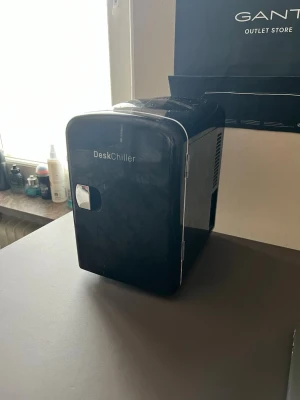 Mini kyl - Snyggt och kompakt sminkkylskåp från DeskChiller i svart plast med vita detaljer. Perfekt för att hålla dina skönhetsprodukter svala och fräscha. Kylskåpet har en modern, rundad design och öppnas med ett handtag i silverfärg. Drivs med sladd.