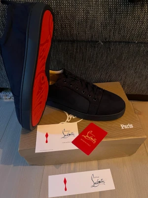 Svarta Christian Louboutin sneakers - Säljer ett par svarta sneakers från Christian Louboutin med klassisk röd sula. Skorna har lågt skaft, svarta skosnören och diskreta metalldetaljer. Tillverkade i mocka för en lyxig känsla och stilren look. Perfekta för dig som vill sticka ut med exklusiva detaljer.