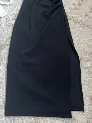 Svart långkjol med slits - Stilren svart tub klänning med slits på sidan. Klänningen har en formande passform och är tillverkad i ett mjukt, följsamt material som ger en snygg siluett. Perfekt för dig som gillar minimalistisk och tidlös stil.