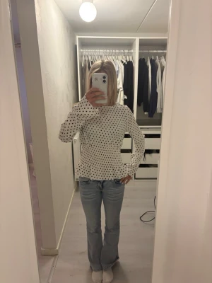 Vit prickig blus med volang - Snygg vit blus med svarta prickar och volang nedtill. Blusen har rund halsringning och långa ärmar. Materialet ser ut att vara mjukt och stretchigt, perfekt för en trendig och bekväm look. Passar till både jeans  och kjol.  Den är för stor för mig därför den är så lång!