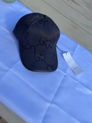 Svart keps med GG-mönster - Snygg svart keps med det klassiska GG-monogrammet över hela kepsen. Kepsen har böjd skärm och justerbar baksida. Tillverkad i ett slitstarkt tyg med exklusiv känsla. Perfekt för dig som vill ha en stilren accessoar med lyxig touch.