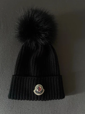 Svart Moncler mössa med pälsboll - Säljer en svart ribbstickad mössa från Moncler med stor fluffig pälsboll på toppen och Moncler-logga framtill. Mössan är tillverkad i Italien och har en uppvikt kant. Perfekt för dig som vill ha en snygg och trendig accessoar till vintern.