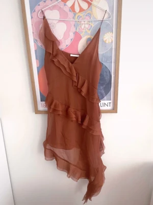 asymmetrisk volangklänning - Säljer en brun klänning med omlott och volangdetaljer. Klänningen har smala axelband, v-ringning och asymmetrisk nederkant som ger en unik look. Perfekt för dig som vill sticka ut med en trendig och luftig stil.
