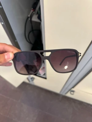 Bruna Tom Ford solglasögon - Snygga svarta solglasögon från Tom Ford med klassisk pilotform och mörka glas. Bågarna har en matt finish och guldiga detaljer vid gångjärnen. Perfekt accessoar för dig som vill ha en stilren och trendig look.