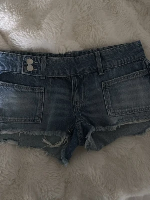 Blå jeansshorts - Säljer ett par blå jeansshorts med hög midja, råa och slitna kanter samt stora framfickor. Shortsen har dubbla silverknappar och bälteshällor. Perfekta för sommaren och ger en avslappnad streetstyle-look.