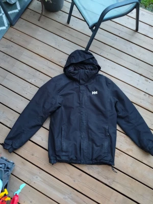 Svart regnjacka från Helly Hansen - Snygg svart regnjacka från Helly Hansen med huva och dragkedja framtill. Jackan har två fickor med dragkedja och justerbar nederkant. Perfekt för regniga dagar och enkel att matcha med olika outfits.