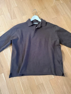 Brun pikétröja med krage - Klassisk brun långärmad pikétröja från Zara . Tröjan har krage, knäppning med två knappar och slitsar i sidorna. Tillverkad i ett mjukt bomullstyg som känns skönt mot huden. Perfekt för en avslappnad men stilren look.