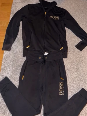Svart hoodie från BOSS med dragkedja - Svart hoodie från BOSS med dragkedja framtill och gulddetaljer på dragkedjorna. Märkets logga syns på bröstet. Hoodien har huva och två fickor med dragkedja. Perfekt för en avslappnad och stilren look. Lång byxor och tröja  stor i tröjan 