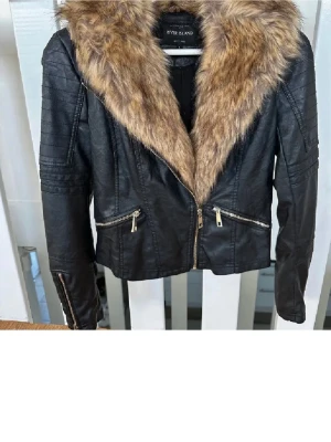 Svart skinnjacka River Island päls - Snygg svart skinnjacka från River Island med stor, fluffig fuskpäls i beige/bruna toner runt kragen. Jackan har dragkedja framtill, två snedställda fickor med dragkedja och detaljerade sömmar på ärmarna. Perfekt för dig som vill sticka ut med en trendig look.