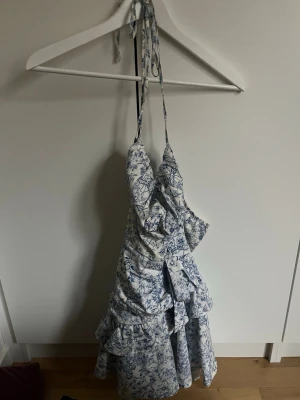 Blommig volangklänning med knyt - Söt sommarklänning med blått blommönster på vit botten. Klänningen har tunna axelband med knyt i nacken, volangdetaljer och omlottkänsla. Perfekt för varma dagar och har en romantisk vibe.