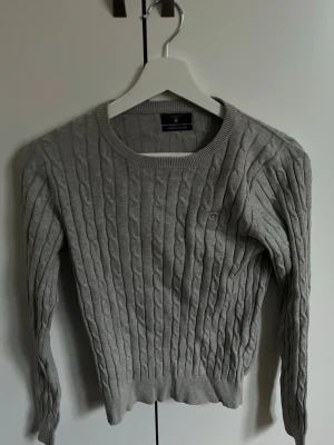 Grå kabelstickad tröja från Gant - Snygg grå kabelstickad tröja från Gant i stretchig bomull. Tröjan har rund halsringning, ribbade muddar och en liten Gant-logga broderad på bröstet. Perfekt för lager på lager och enkel att matcha med jeans eller kjol.