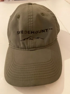 Olivgrön keps från Swedemount - Snygg olivgrön keps från Swedemount med böjd skärm och broderad logga framtill. Klassisk sexpanelsdesign med ventilationshål och justerbar passform bak. Tillverkad i slitstarkt tyg, perfekt för en aktiv livsstil.
