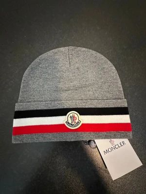 Grå Moncler mössa med ränder - Snygg grå mössa Moncler med klassiska svarta, vita och röda ränder på uppviket. Mössan har Moncler-logga framtill och är tillverkad i mjukt material som känns skönt mot huden. Perfekt accessoar för att lyfta din streetwear-look.
