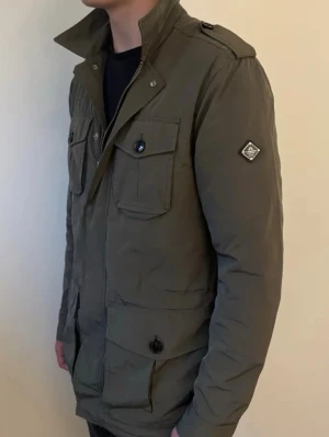 Mörkgrön J Lindeberg Field Jacket - Snygg olivgrön parkasjacka med fyra stora fickor framtill och axelklaffar. Jackan har hög krage, dragkedja och knappar samt en patch på ena ärmen. Perfekt för dig som gillar en stilren och funktionell look. Storlek M, nytt skick använd väldigt få gånger, passar dig runt 185.