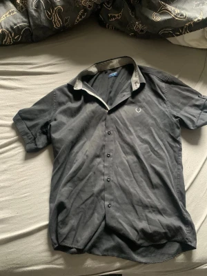 Svart kortärmad skjorta Fred Perry - Snygg svart skjorta från Fred Perry med korta ärmar och klassisk krage. Skjortan har knappar framtill och ett diskret logobroderi på bröstet. Insidan av kragen har ett rutigt mönster i beige och svart. Perfekt för en stilren look.