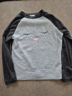 Tröja - Supermjuk och mysig tröja i grått med lång ärm. Framsidan är i fluffigt teddy-material med ett sött broderat kattansikte och texten 'SLEEP ALL DAY' längst ner. Ärmar och baksida är i mörkare grått tyg.
