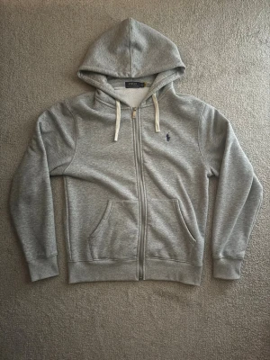 Grå zip hoodie från Polo Ralph Lauren - Snygg grå hoodie med dragkedja från Polo Ralph Lauren. Klassisk design med huva, vita dragsnören och två fickor framtill. Broderad marinblå logga på bröstet. Tillverkad i mjukt sweatshirtmaterial som är perfekt för chill dagar. Liten i storlek.