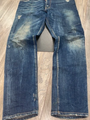 Blå jeans från Dsquared2 - Säljer ett par blå jeans från Dsquared2 med slitna detaljer och kontrastsömmar. Jeansen har klassisk femficksmodell och en röd logotypetikett vid gylfen. Materialet är denim i bomull och passformen är rak.