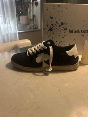 Mörk blåa  sneakers från The Gallerist - Snygga svarta sneakers från The Gallerist med vit klöverdetalj på sidan, vita skosnören och vit häl. Skorna har en klassisk låg siluett och är tillverkade i mockaliknande material. Perfekta för dig som vill ha en unik touch på din outfit.
