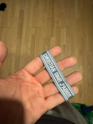 Blått Christian Dior J'Adior armband - Snyggt blått och vitt tygarmband från Christian Dior med texten 'J'Adior'. Armbandet har en vävd design och avslutas med tofsar i ändarna. Perfekt accessoar för dig som vill ha en exklusiv och trendig detalj till din outfit.