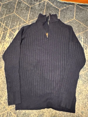 Mörkblå stickad tröja med halvzip - Mörkblå stickad tröja med ståkrage och halv dragkedja framtill. Tröjan har ett ribbat mönster längs hela plagget och långa ärmar. Perfekt för lager-på-lager och en stilren look.