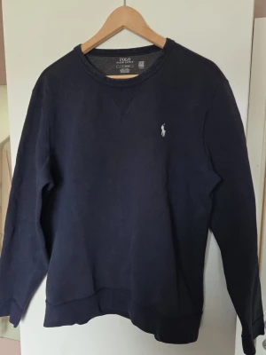 Mörkblå sweatshirt från Polo Ralph Lauren - Klassisk mörkblå sweatshirt från Polo Ralph Lauren med rund halsringning och den ikoniska vita logotypen broderad på bröstet. Tröjan har långa ärmar och ribbade muddar vid ärmslut och nederkant. Perfekt för en avslappnad och stilren look. OBS något urtvättad vid halsen men i övrigt väldigt bra skick.