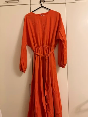 Lång orange klänning med bälte - Säljer en lång, orange klänning med rund halsringning och långa ärmar. Klänningen har ett matchande bälte i midjan och stängs med en knapp i nacken. Perfekt för dig som gillar färgstarka plagg och enkel, stilren design.
