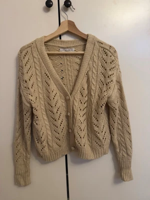 stickad kofta - Superfin beige stickad kofta från NA-KD med v-ringning och dekorativa pärlknappar. Koftan har ett snyggt hålmönster och kabelstickade detaljer, samt ribbade muddar vid ärmslut och nederkant. 