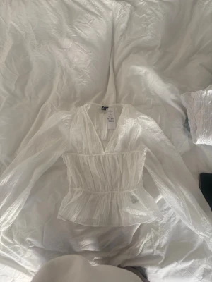 Vit blus med dragsko från Ginatricot - Superfin vit blus från Ginatricot med dragsko i midjan och v-ringning. Blusen har långärmade ärmar och är gjord i ett lätt, transparent tyg med struktur. Perfekt att styla med jeans eller kjol för en trendig look.