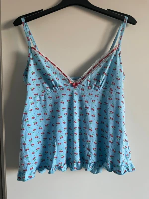 Blå linnetopp med körsbär och spets - Supersöt blå linnetopp med tunna axelband och körsbärsmönster över hela tyget. Toppen har vit spetskant vid urringningen och en liten rosett framtill. Volangkant nertill ger en extra gullig touch. Perfekt för chill dagar hemma eller sommarhäng.