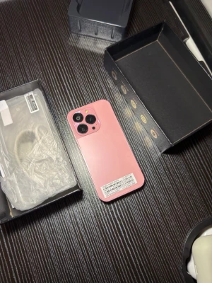 Rosa iPhone med dubbla kameror - Säljer en snygg rosa iPhone med stilren design och rundade kanter. Baksidan har två stora kameralinser och en blixt. Telefonen är tillverkad i glas och metall och har en modern look som sticker ut. Perfekt för dig som vill ha en färgglad och trendig mobil.