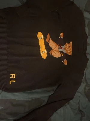 Mörkblå polo bear tröja  - Stickad mörkblå tröja från Ralph Lauren med broderad björn som åker skateboard på framsidan. RL-bokstäver i gult nere vid fållen. Långa ärmar och ribbade muddar vid ärmslut och nederkant. Perfekt för dig som gillar streetstyle och unika detaljer.