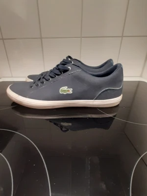 Mörkblå Lacoste sneakers - Snygga mörkblå sneakers från Lacoste med klassisk låg profil och vitt gummisula. Skorna har det ikoniska gröna krokodilmärket på sidan och är tillverkade i textil och syntetmaterial. Perfekta för en stilren och sportig look. Storlek 41.