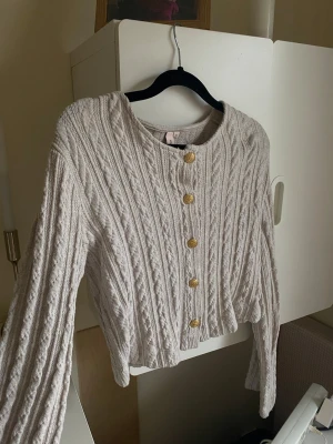 Beige kabelstickad kofta med guldknappar - Trendig beige kabelstickad kofta med rund hals och snygga guldknappar framtill. Kortare modell med långa ärmar och ribbade muddar. Perfekt att styla till jeans eller kjol❤️