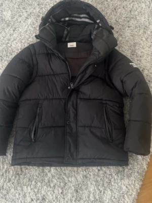 Svart pufferjacka från Burberry - Säljer en svart pufferjacka från Burberry London England med huva och dragkedja framtill. Jackan har två framfickor med dragkedja och en tydlig Burberry-logga på ärmen. Perfekt för kalla vinterdagar och riktigt snygg streetstil.