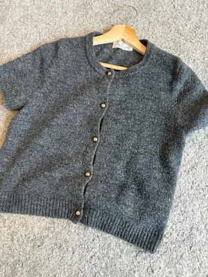 Mörkgrå kortärmad kofta med knappar - Säljer en mörkgrå stickad kofta med korta ärmar och rund hals. Koftan har guldfärgade knappar framtill och ribbade kanter. Perfekt att slänga över en t-shirt för en chill look.