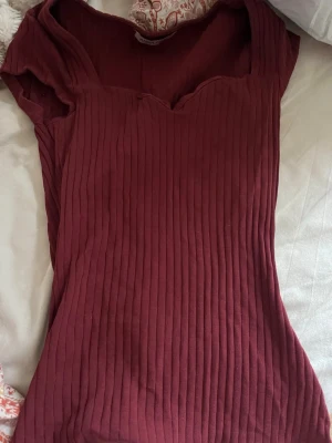 Ribbad vinröd topp  - Snygg vinröd ribbad topp med korta ärmar och en unik sweetheart-ringning. Toppen har en figurnära passform och är tillverkad i mjuk bomullsmix som känns skön mot huden. Perfekt att styla med jeans eller kjol för en trendig look.