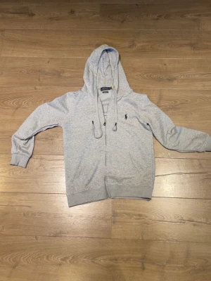Grå hoodie från Polo Ralph Lauren - Snygg ljusgrå hoodie från Polo Ralph Lauren med dragkedja framtill och justerbar huva. Klassisk broderad logga på bröstet och ribbade muddar vid ärmar och nederkant. Tillverkad i mjukt bomullsmaterial för extra komfort.
