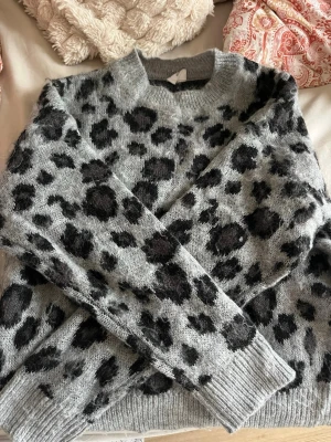 Grå leopardmönstrad stickad tröja - Säljer en grå stickad tröja från & Other Stories i storlek XS. Tröjan har ett coolt svart leopardmönster, rund halsringning och ribbade muddar vid ärmslut och nederkant. Perfekt för dig som vill ha en trendig och mysig look.